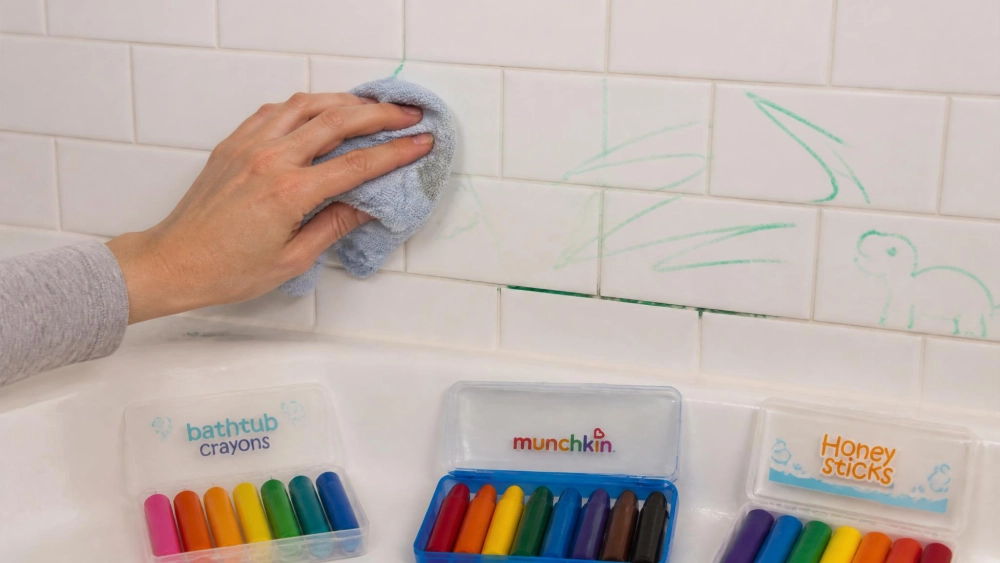 A parent's hand wiping green bathtub-marker lines off a white tile wall with a damp cloth, a stubborn streak still in the grout line below, and several open bath crayon sets on the tub edge.