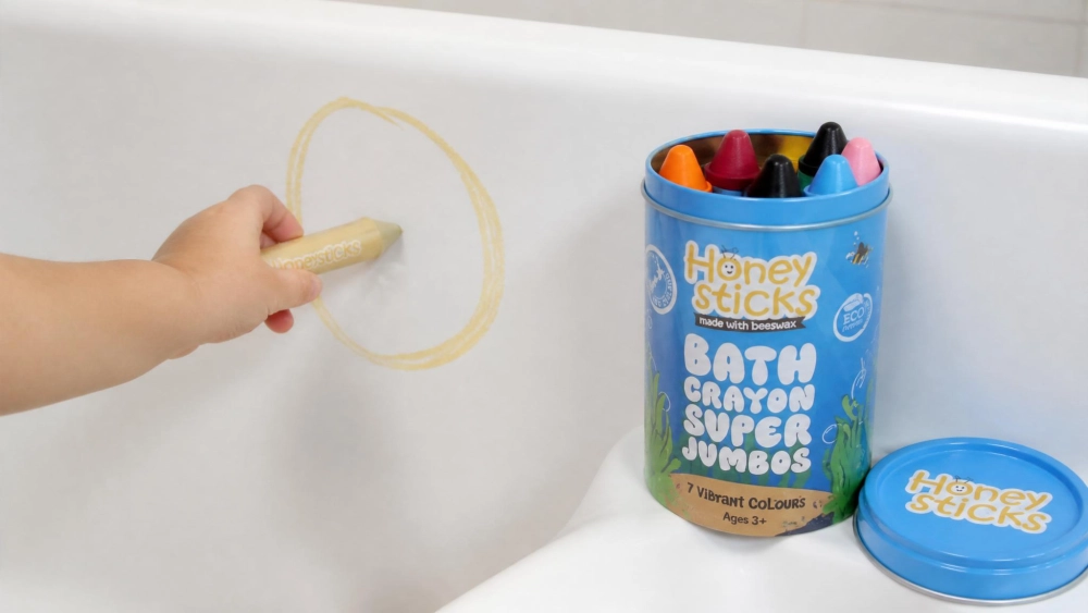 A toddler's hand gripping a thick natural-colored Honeysticks beeswax bath crayon and drawing a wobbly circle on a white acrylic tub wall, with the storage tin of jumbo crayons beside the tub.