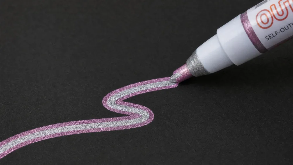 A close-up of a self-outline shimmer marker drawing a two-tone metallic line on black paper, showing the colored outline around a silver center.