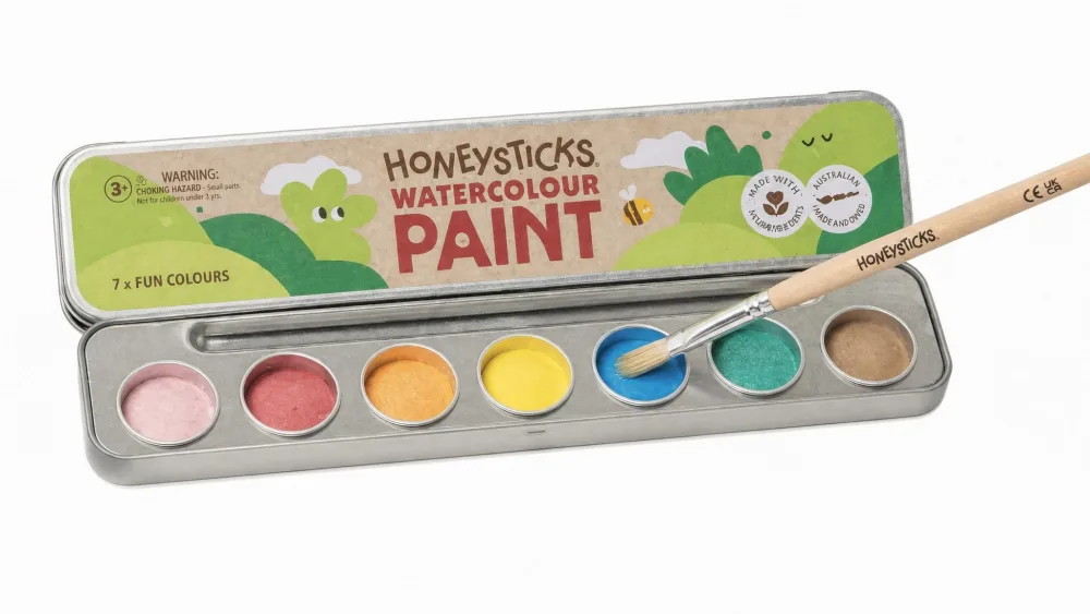 The Honeysticks 8-color watercolor palette open in its metal tin, a wet brush touching one of the solid color pans.