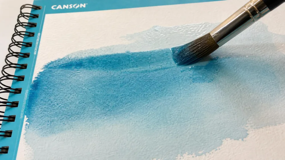 A paintbrush spreading blue watercolor paint across damp Canson XL paper