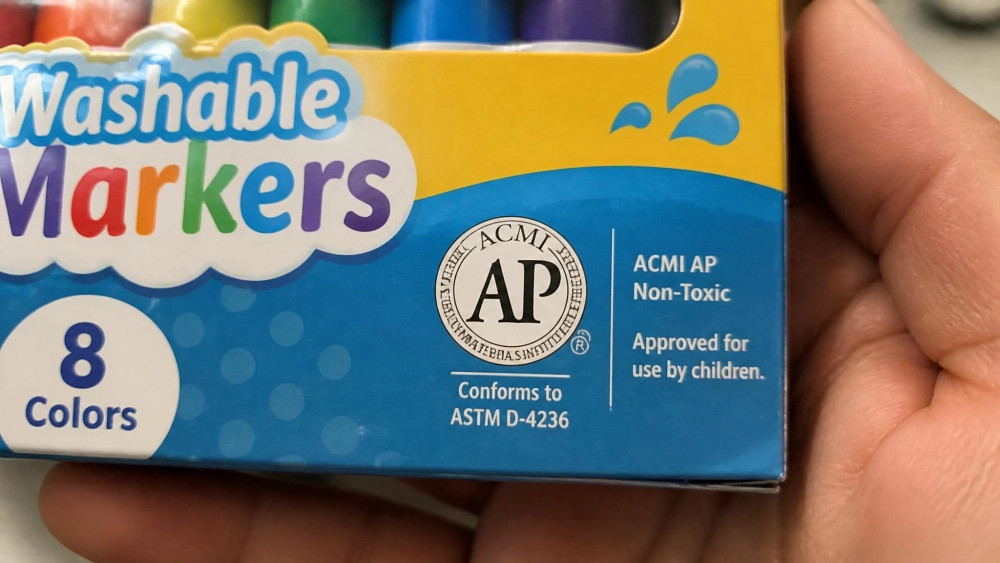Close-up of a kids' marker package showing the ACMI AP non-toxic seal and an ASTM D-4236 conformance line, held in an adult's hand.