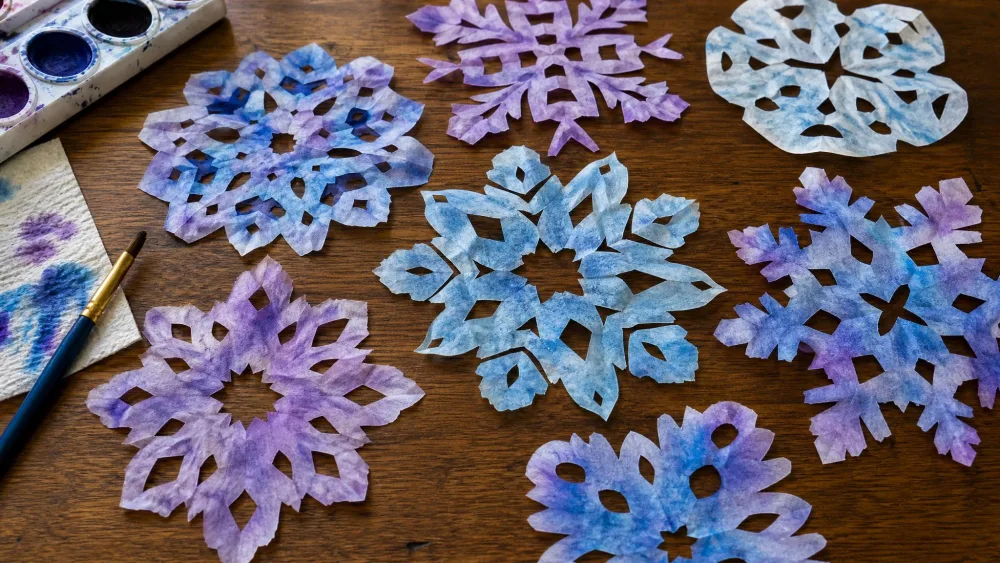 Coffee filters cut into snowflakes and painted with blue and purple watercolor, drying on a table.