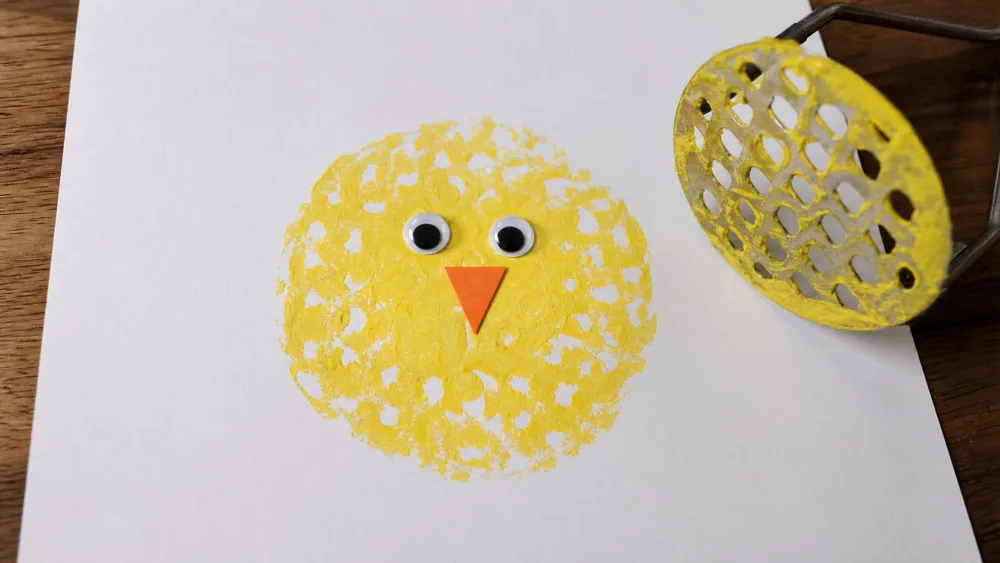A round yellow chick print stamped with a potato masher, with an orange triangle beak and googly eyes added, drying on white paper.