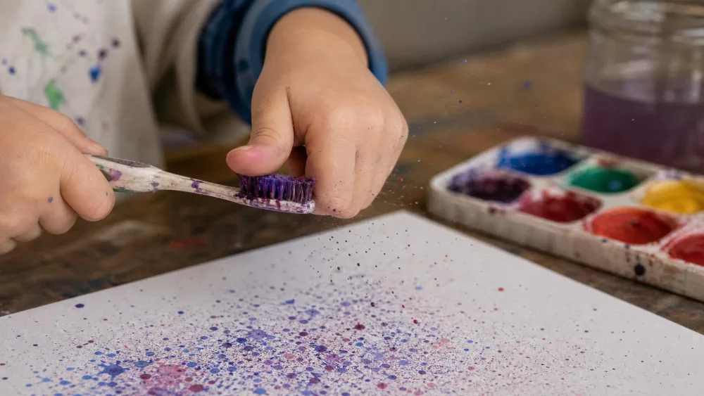 A child's hand running a thumb across an old toothbrush loaded with thinned watercolor paint, flicking fine splatters onto a sheet of paper below.