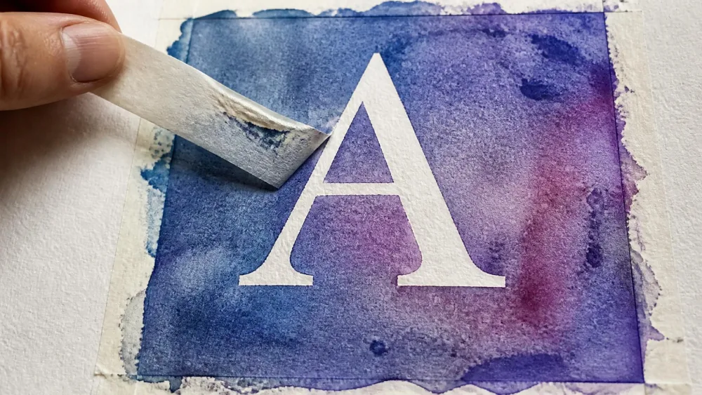 Tape peeled from dried watercolor revealing a white initial.