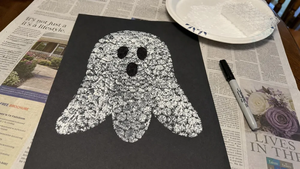 A white dotted ghost shape printed with bubble wrap onto black paper, with marker eyes, drying on a newspaper-covered kitchen table.