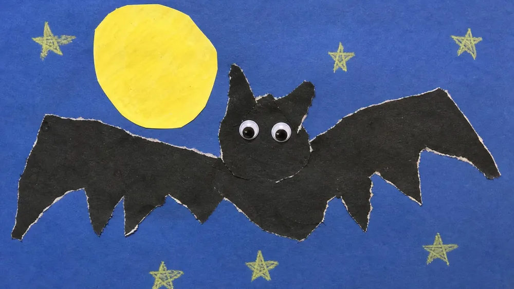 A torn black paper bat with jagged wings and googly eyes glued onto a paper night sky with a yellow full moon, made by a young child.