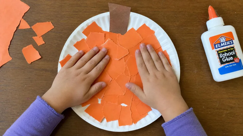 A child's hands pressing torn orange construction paper onto a paper plate to make a pumpkin, with a brown paper stem at the top and a bottle of white glue nearby.