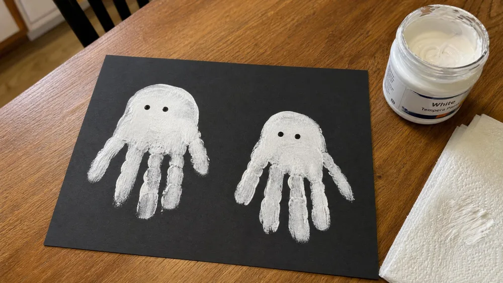 Two white painted handprint ghosts on black construction paper with small dot eyes, drying on a kitchen table beside a jar of white paint.