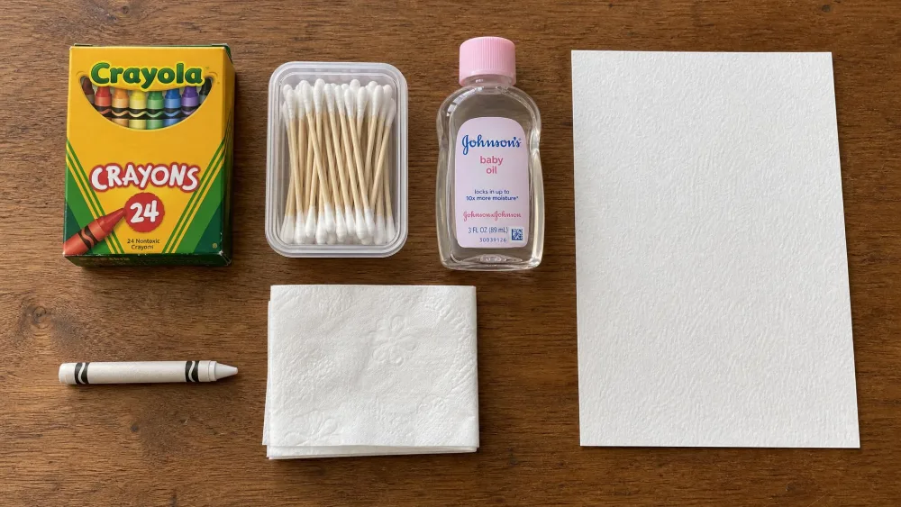 A flat-lay on a wooden kitchen table showing a box of crayons, cotton swabs, a small bottle of baby oil, a white crayon, a folded tissue, and a sheet of textured drawing paper.