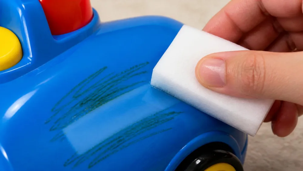 A hand holding a damp white melamine foam eraser wiping green crayon off a glossy blue plastic toy, showing a clean streak where the mark has lifted.