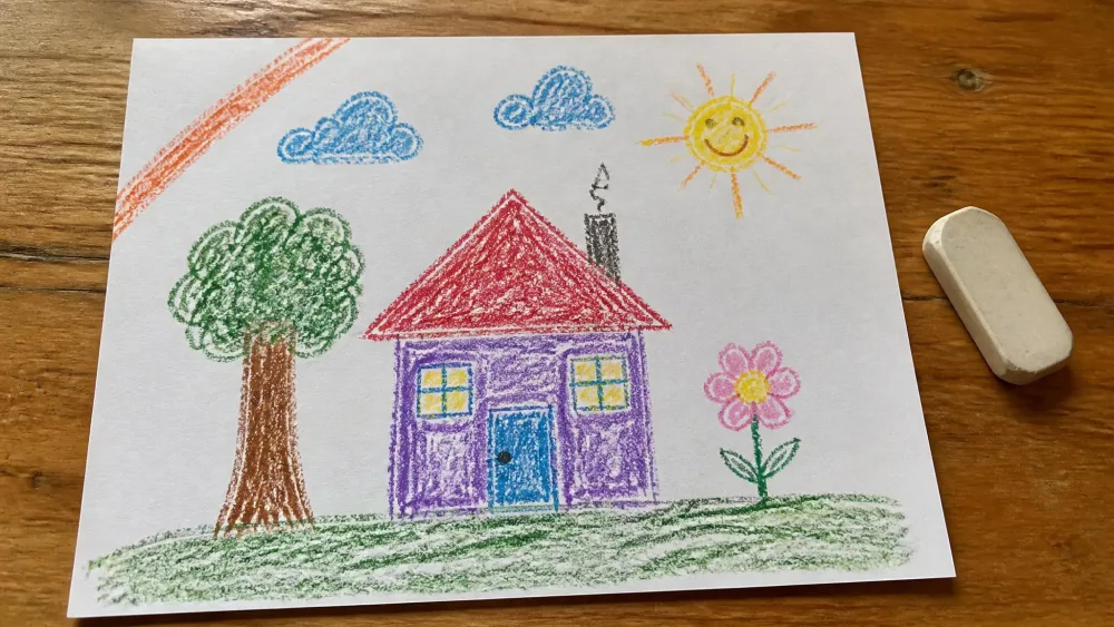A child's crayon drawing on white paper on a wooden kitchen table, with a stray orange crayon streak across one corner and a soft eraser resting beside the page.