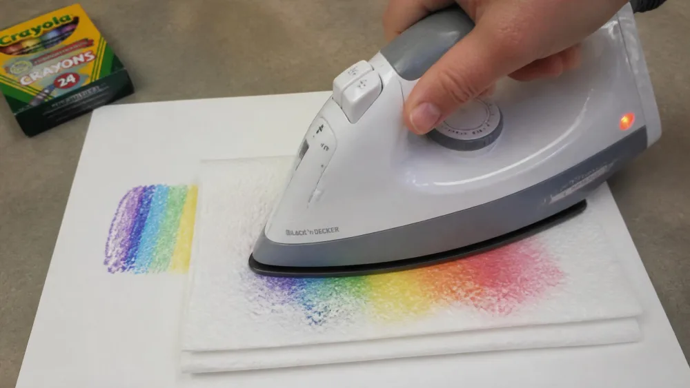 An adult pressing a low-setting iron onto folded white paper towels laid over a crayon mark on sturdy paper, with melted wax visibly soaking into the towel.