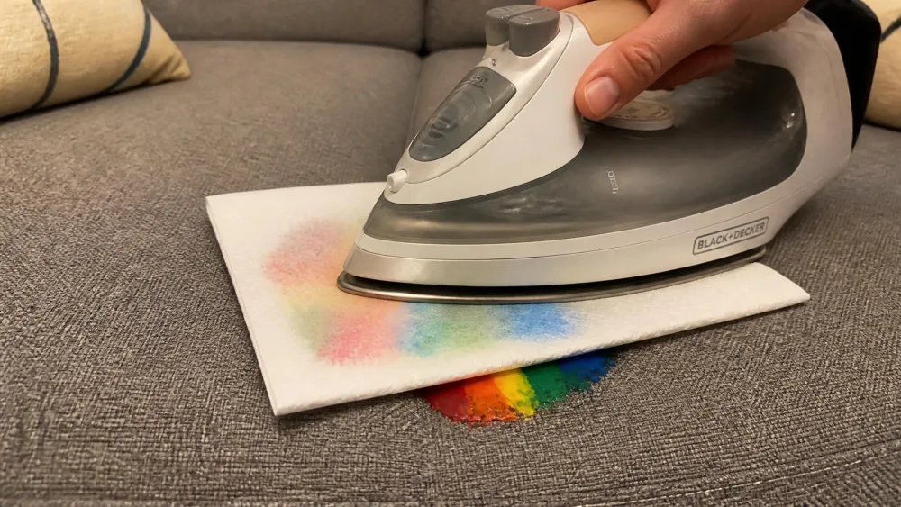 A warm iron pressing a folded white paper towel over a melted crayon spot on a gray upholstered couch, with wax visibly soaking into the paper.