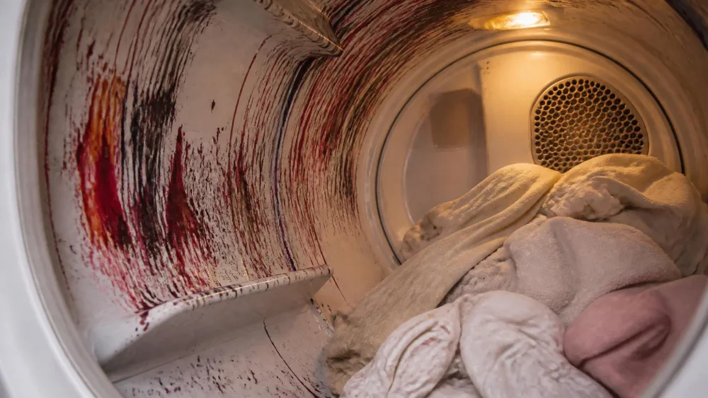 The inside of a white dryer drum streaked with dark red and black melted crayon wax, a warm load of light-colored laundry pushed to one side.