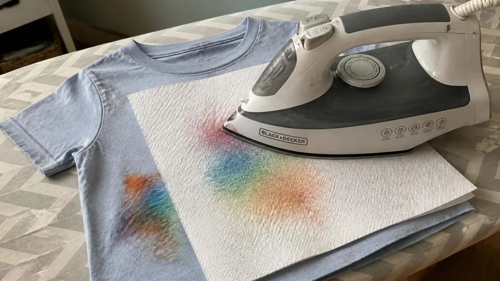 An iron pressing on a white paper towel laid over a stained child's t-shirt on an ironing board, with a colored waxy crayon blot visibly transferred onto the paper towel underneath.