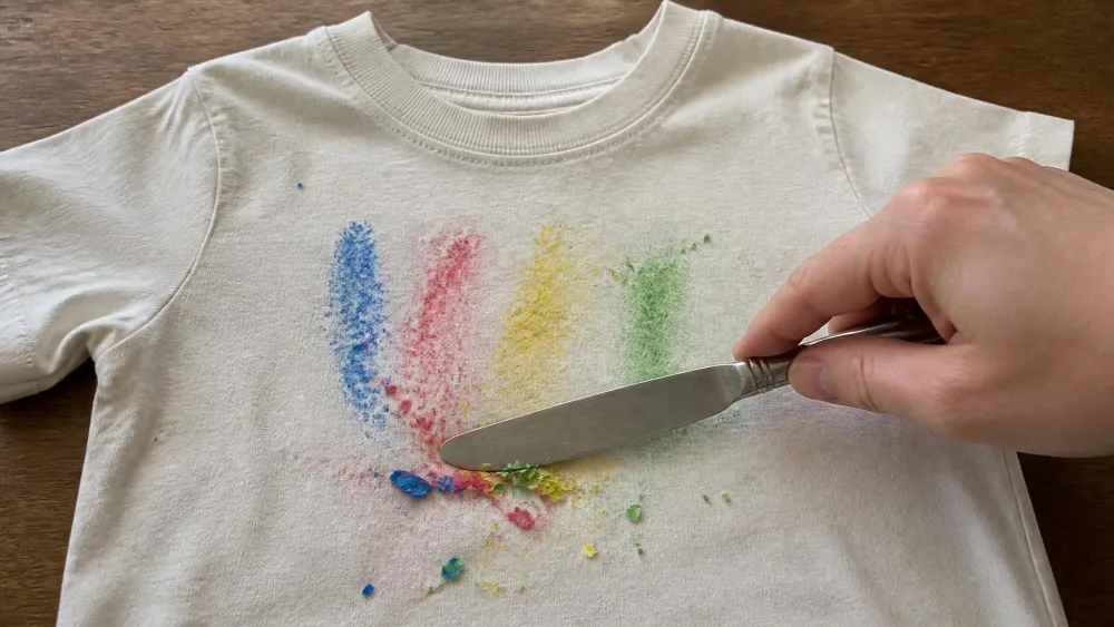 A dull butter knife scraping brittle, frozen crayon flakes off the surface of a child's cotton t-shirt laid flat on a table.