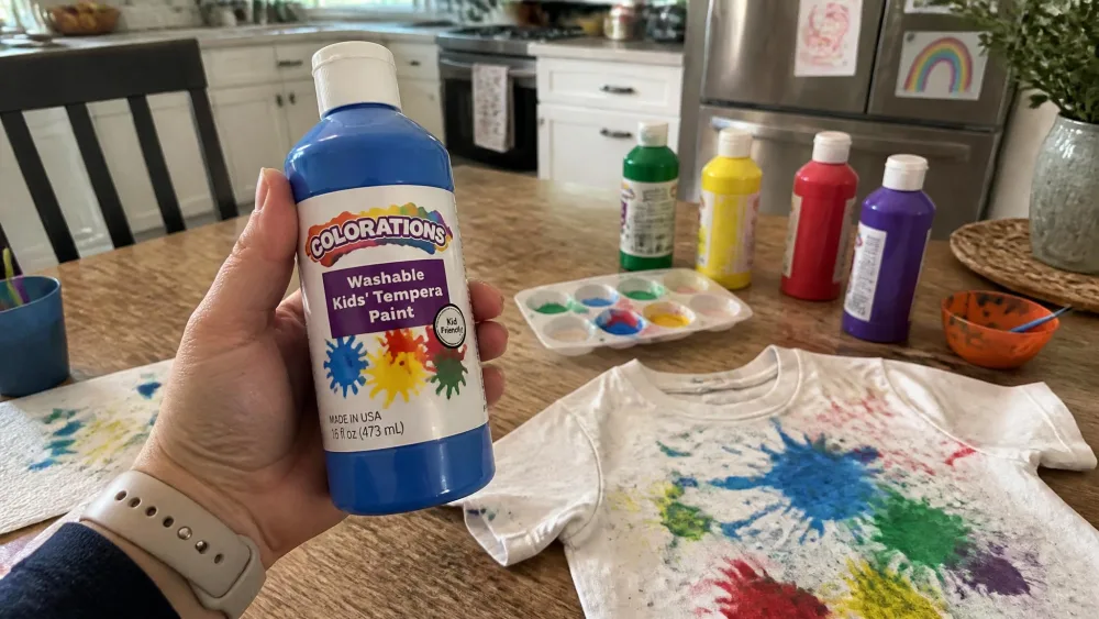A parent's hand holding a bottle of washable kids' tempera paint label-side up over a kitchen table scattered with paint bottles and a stained child's t-shirt.