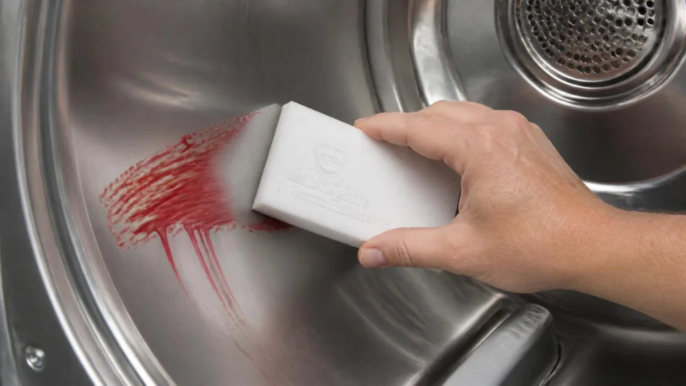 A hand using a white Mr. Clean Magic Eraser to scrub a streak of melted red crayon off the stainless steel interior drum of a clothes dryer.