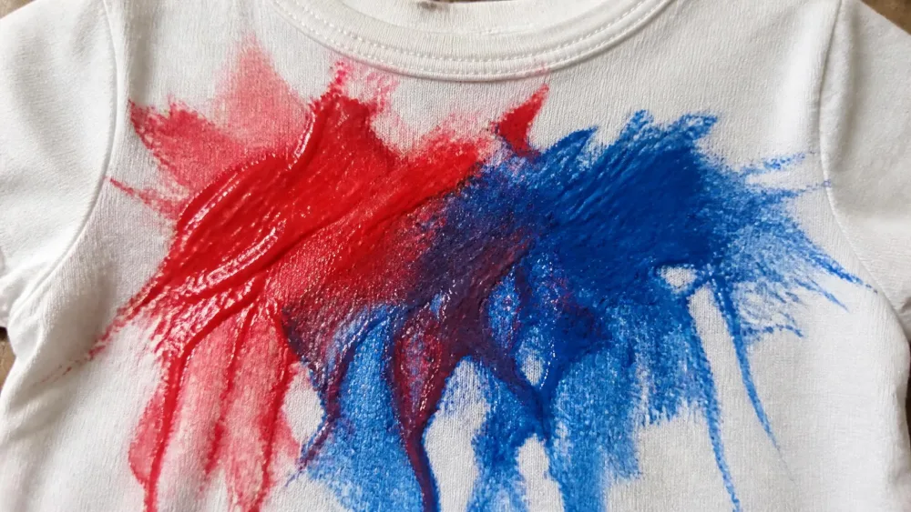 Close-up of a red and blue melted crayon stain smeared across the front of a white cotton child's t-shirt, with the waxy sheen of the crayon visible on the fabric surface.