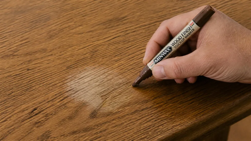 A hand running a wood touch-up marker along the grain of an oak tabletop to blend a dull cleaned spot.