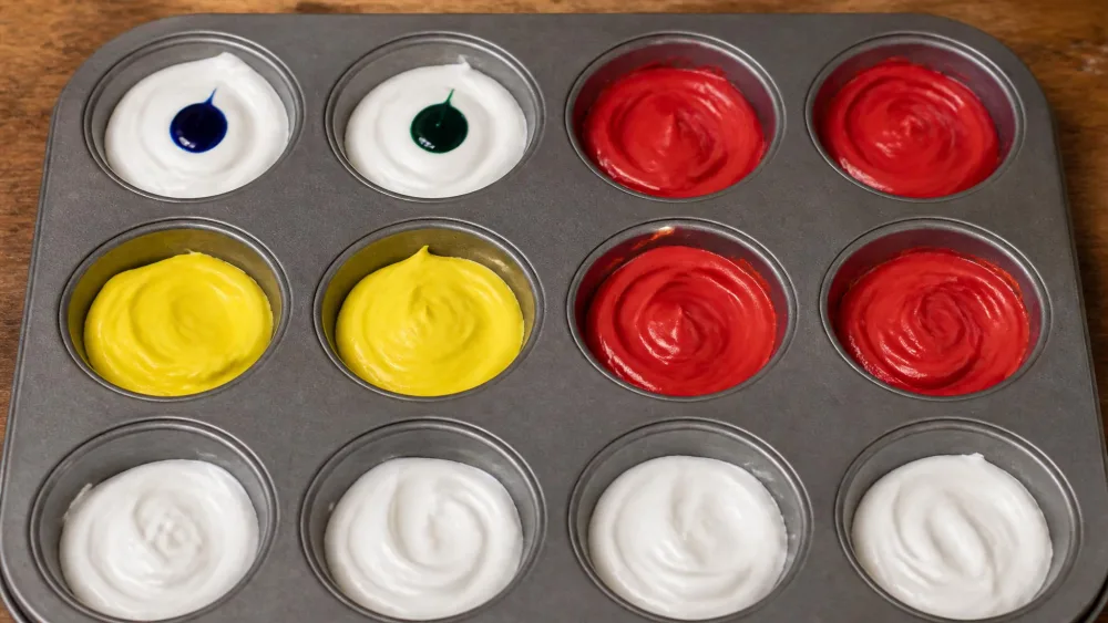 Drops of blue and green food coloring squeezed into separate cups of white paint paste in a mini muffin tin, with several cups already colored red and yellow.