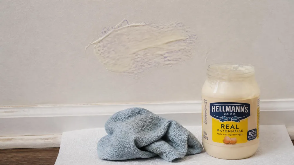 A small smear of full-fat mayonnaise spread over a faded crayon mark on a painted wall, with a soft cloth and a jar of mayonnaise resting on a paper towel beneath it.