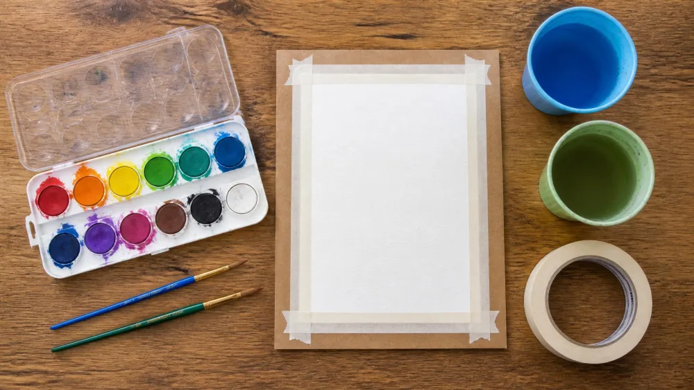 A flat-lay of kids' watercolor supplies on a kitchen table: an open washable pan set, two round brushes, white paper taped to a board, two water cups, and painter's tape.
