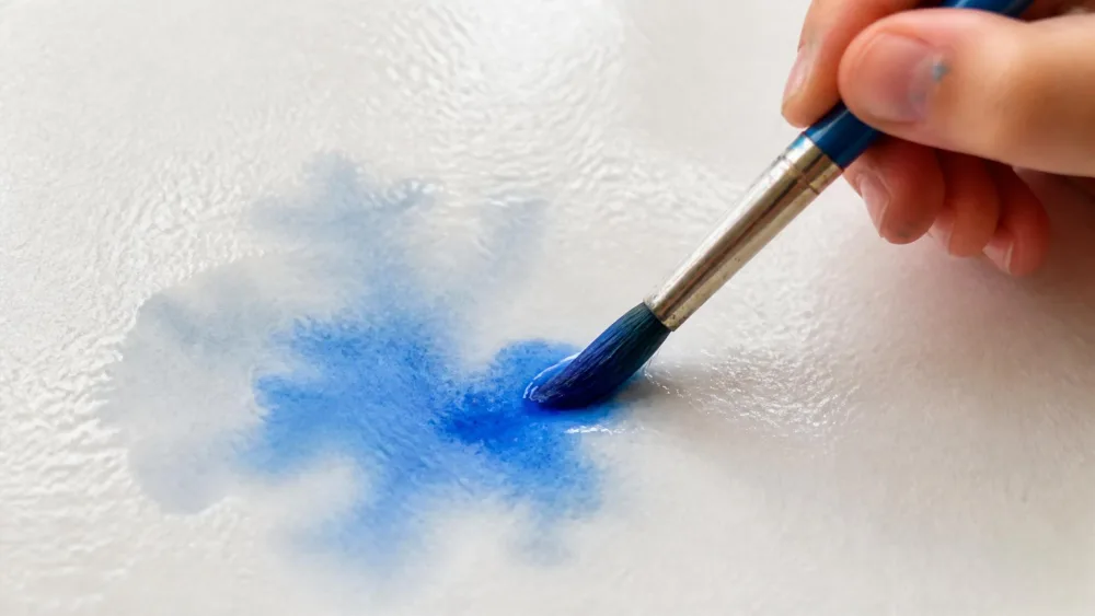 A child's brush touching blue paint onto wet white watercolor paper, the color blooming into a soft cloud where it meets the water.