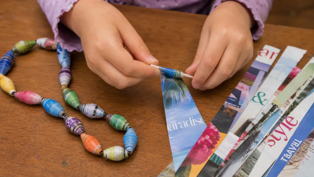 A child's hands rolling a triangle of glossy magazine paper into a bead, with a finished strand of colorful rolled paper beads and cut magazine strips on the table.