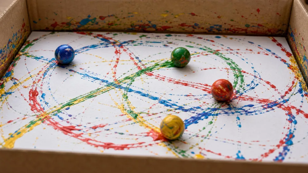 Paint-covered marbles rolling across paper in a shallow box, leaving looping trails.