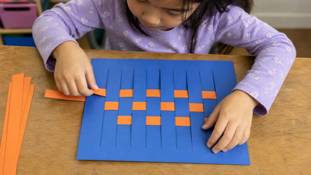 A child weaving orange paper strips over and under slits in a blue sheet to form a woven placemat.