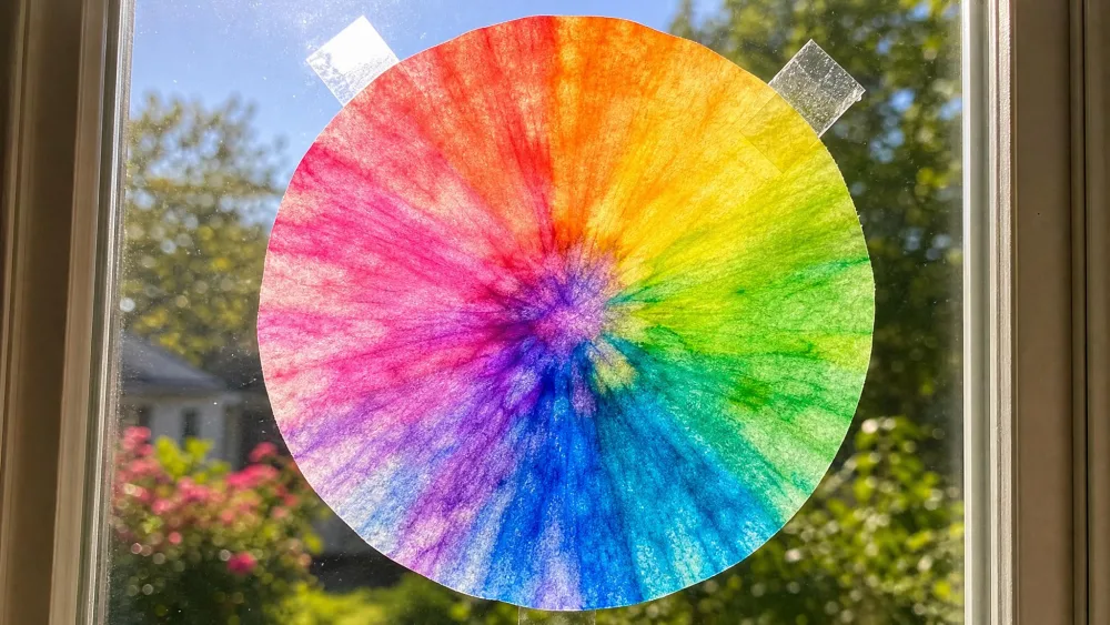 A coffee filter colored with washable markers in bright rainbow tones, taped to a sunlit window and glowing with backlight.