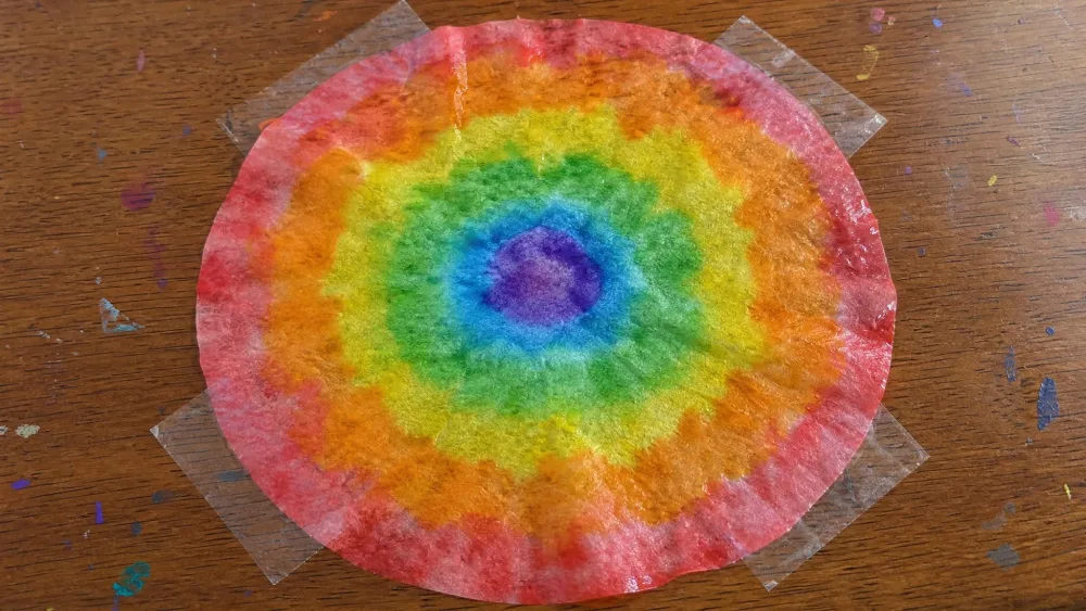 A wet coffee filter taped to a table with concentric rainbow watercolor bands blooming from the center outward, painted by a young child.