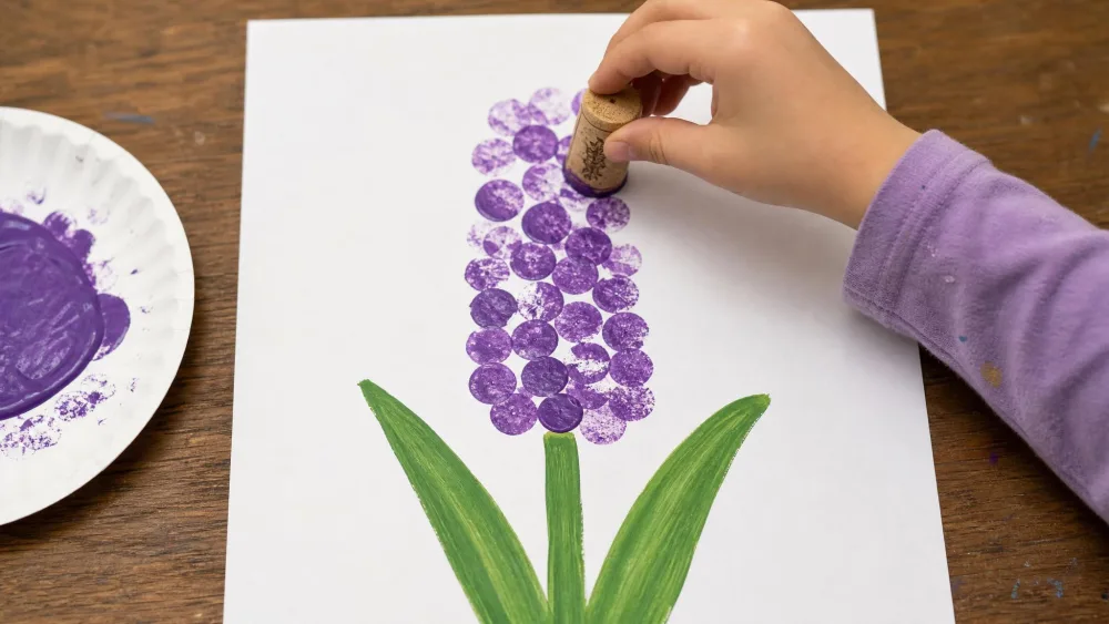 A child stamping purple paint with a wine cork in a vertical column to form a hyacinth flower, with a painted green stem and leaves below.