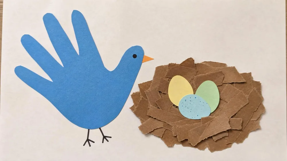 A handprint traced and cut out as a blue bird body, glued beside a nest of torn brown paper strips holding three small paper eggs.