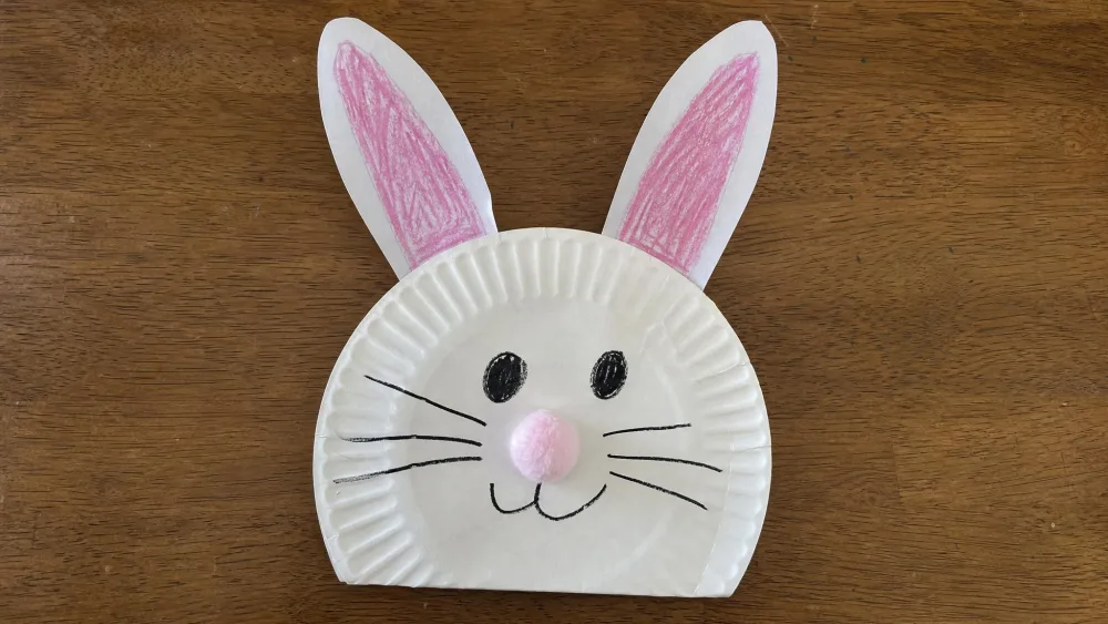 A white paper plate turned into a bunny face with tall pink-lined paper ears, a cotton-ball nose, and drawn whiskers, made by a toddler.