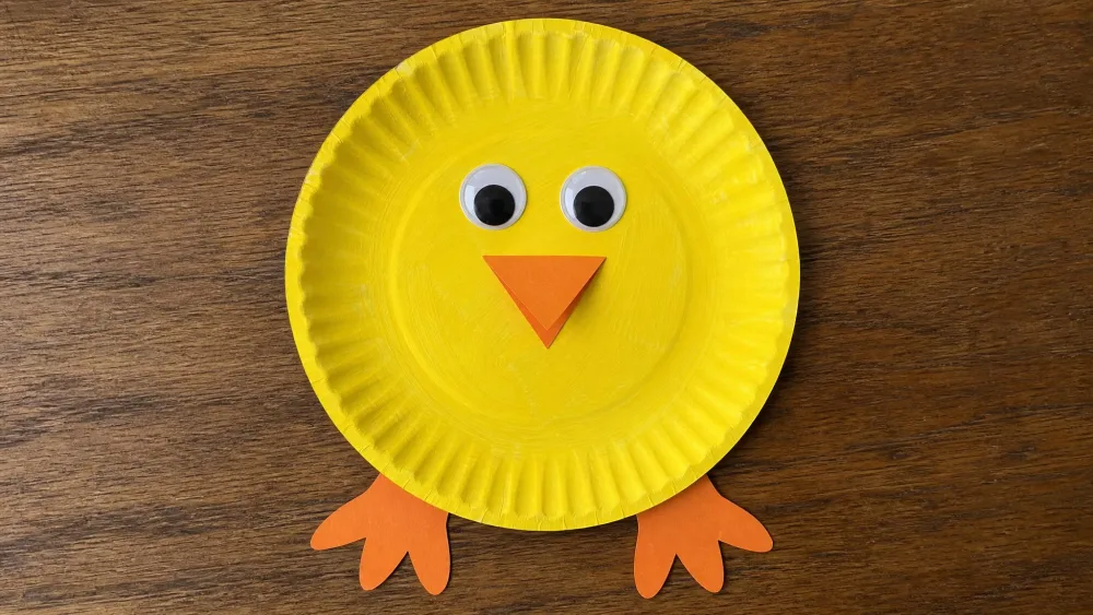 A paper plate painted yellow with an orange construction-paper beak and feet and googly eyes, assembled into a baby chick.