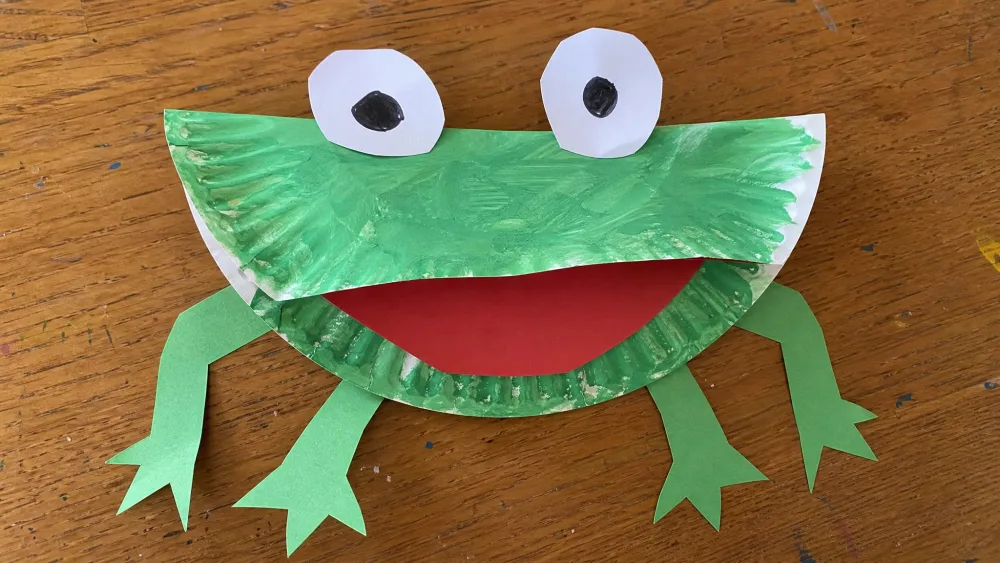 A paper plate painted green and folded in half into a frog with a red paper mouth, white paper eyes, and paper legs, made by a preschooler.