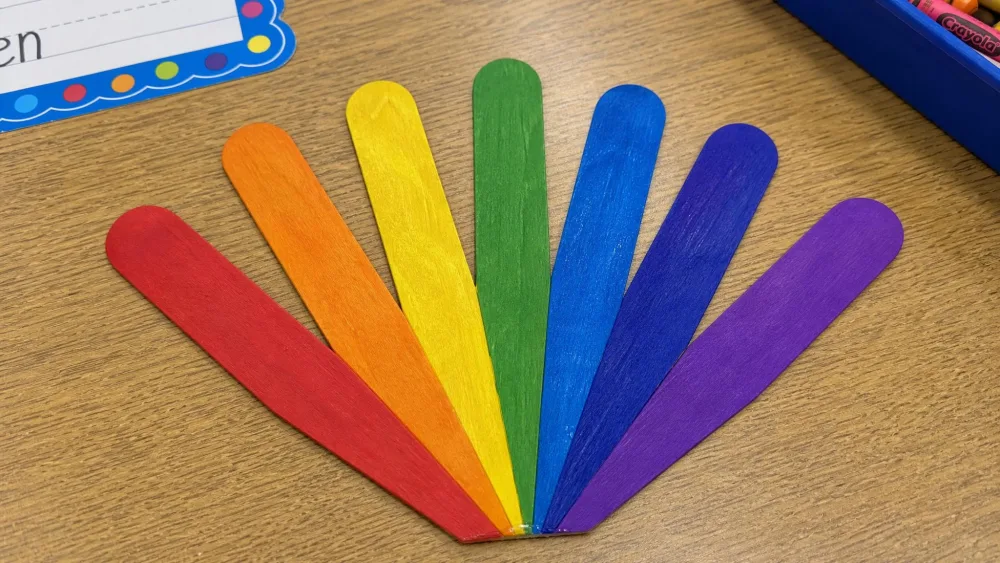 Seven popsicle sticks painted in rainbow colors, glued in a graduated fan shape to form a rainbow, on a classroom table.