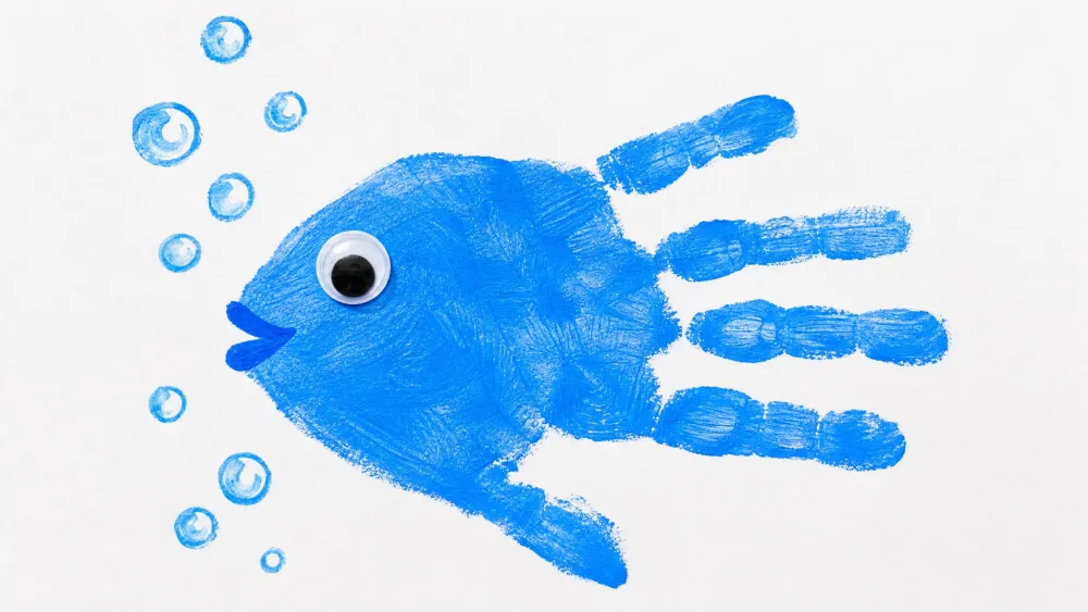A small child's blue-painted handprint pressed sideways on white paper to form a fish with the fingers as the tail, a googly eye and painted water bubbles added around it.