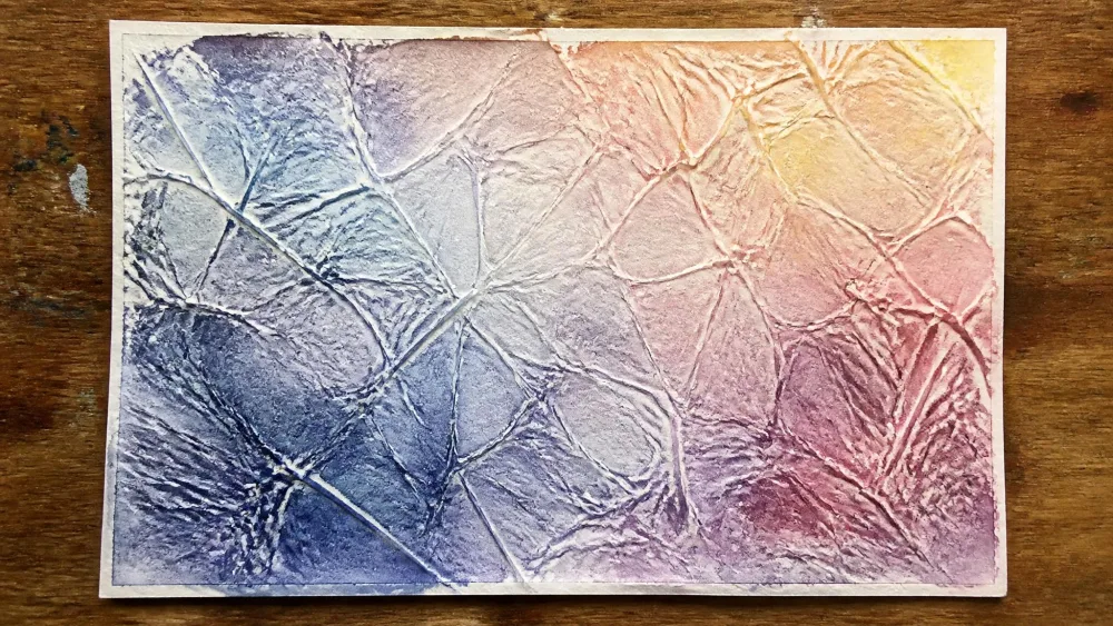 A sheet of plastic cling wrap pressed into a wet watercolor painting, left to dry overnight on a table to create a crackled, geometric texture.