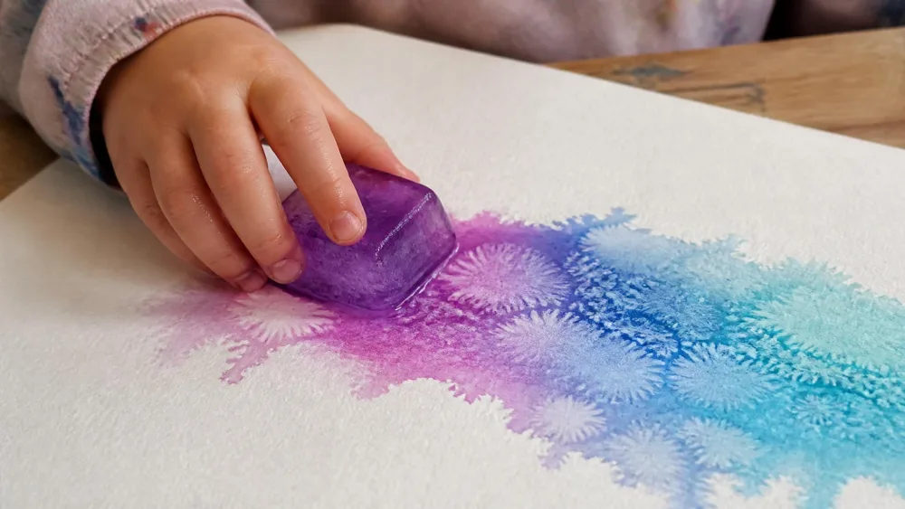 A child's hand sliding a tinted ice chunk across watercolor paper, melted color trailing behind it in an uneven, frosty pattern.