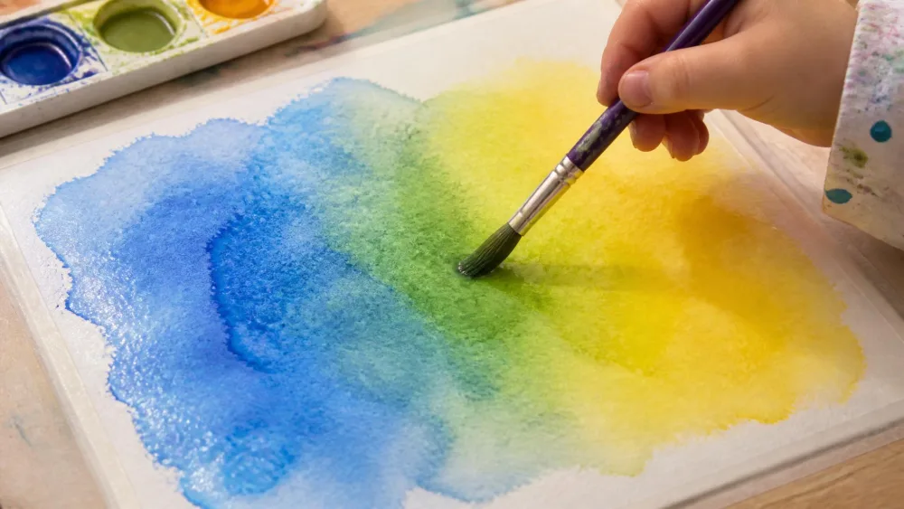 A child's paintbrush blending blue and yellow watercolor on pre-wetted paper, the two colors merging into green at the center of the page.