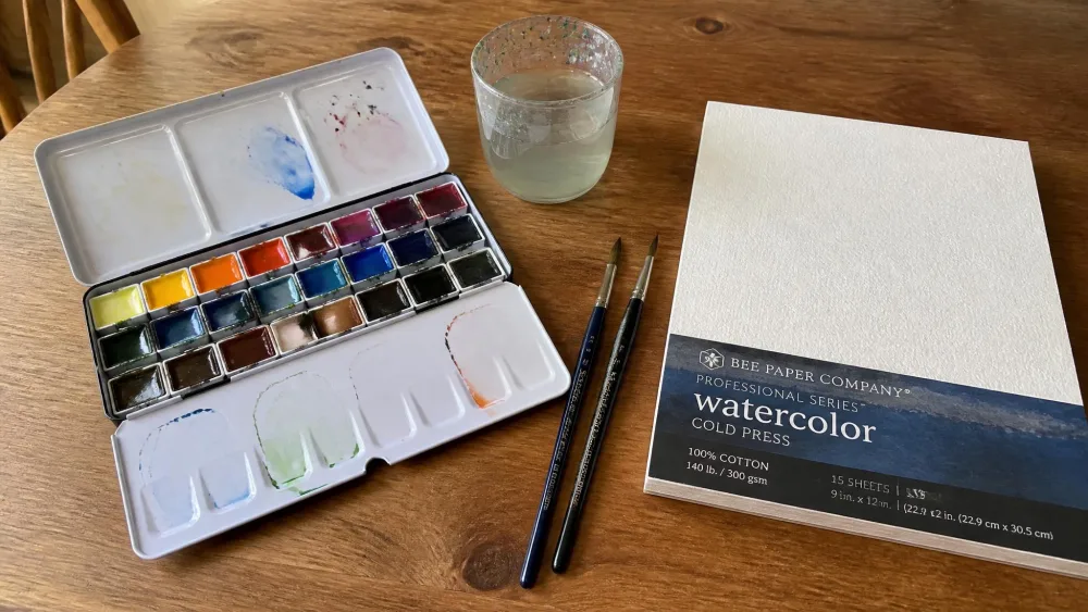 An open pan watercolor set, two round brushes, a water cup, and a pad of cold-press watercolor paper laid out on a wooden kitchen table.