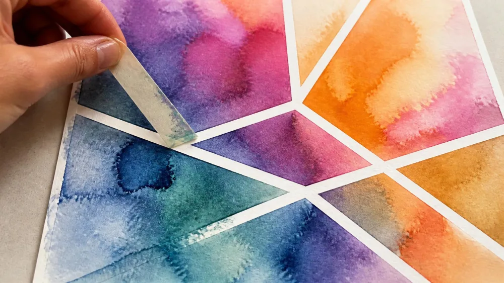 Hand peeling tape off abstract watercolor to reveal white lines.