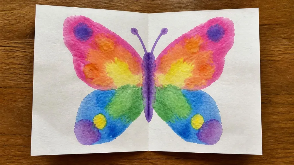 A folded piece of paper opened to reveal a symmetrical butterfly-shaped watercolor blot in bright colors, made by a child.