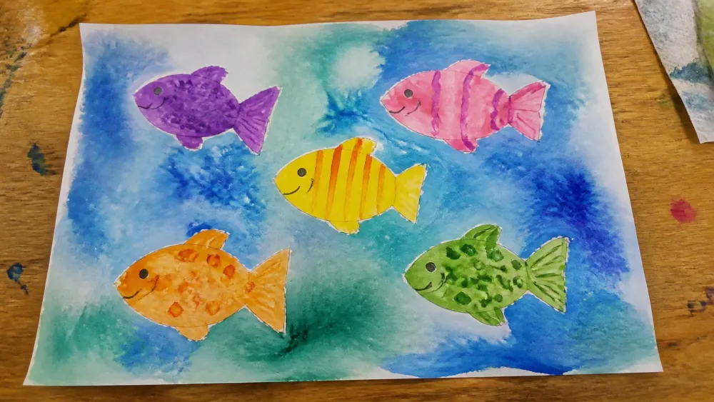 A child's finished watercolor painting of colorful fish shapes against a blue-green wet-on-wet ocean background, drying on a table.