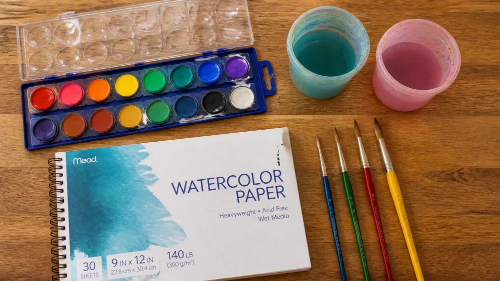 A flat-lay of kids' watercolor supplies on a kitchen table, including a pan of liquid watercolors, a pad of watercolor paper, round paintbrushes, and two rinse cups.